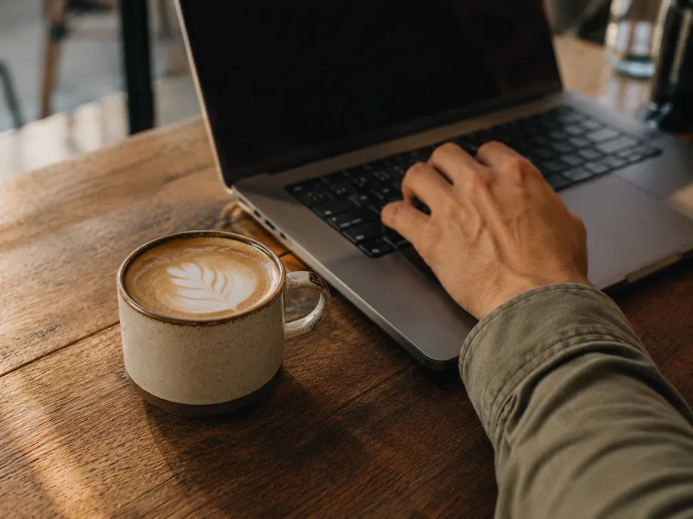 A cortado in a ceramic cup beside an open laptop on a cafe table, a hand on the keys