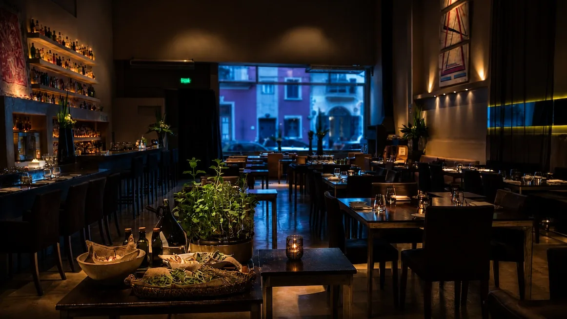 A dim, candlelit dining room with a long backlit wall of bottles, wooden tables set with wine glasses, a potted herb plant on the front table, and a dusk-lit street framed in the back window.