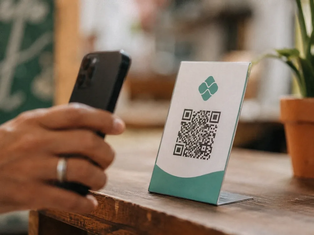 A close view of a hand holding a phone toward a QR-code standee on a wooden café counter