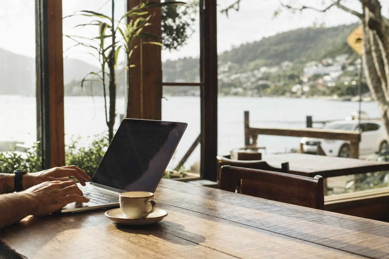 A laptop and a small coffee cup on a wooden table at a cafe near Lagoa da Conceicao, Florianopolis, morning light through the windows
