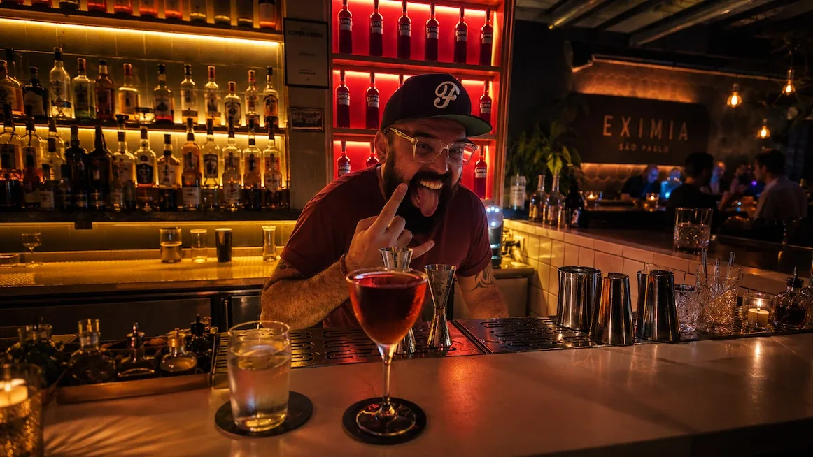 A bartender behind the counter at Exímia — a backlit wall of bottles, jiggers and shakers at hand, a red cocktail and a highball on the bar, and the lit Exímia São Paulo sign glowing on the back wall.