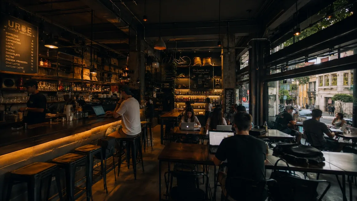 The interior of Urbe Coffee Roasters — a long lit wooden bar with stools, a chalk menu board on the wall, and laptop workers at wooden tables beside big windows onto the street.