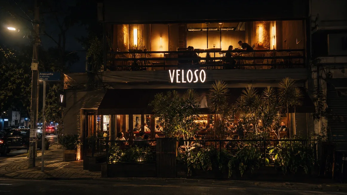 The corner bar Veloso at night, its lit sign above a burgundy awning, an upstairs dining room glowing behind glass, and guests at plant-fringed sidewalk tables.