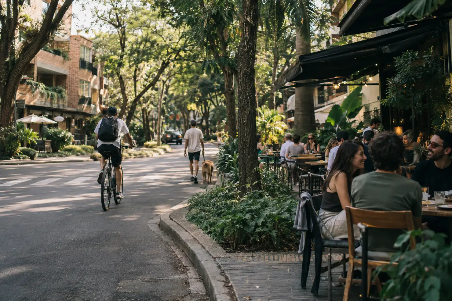A tree-lined street of brick apartment buildings, a cyclist and someone walking a dog passing a sidewalk café underneath the trees