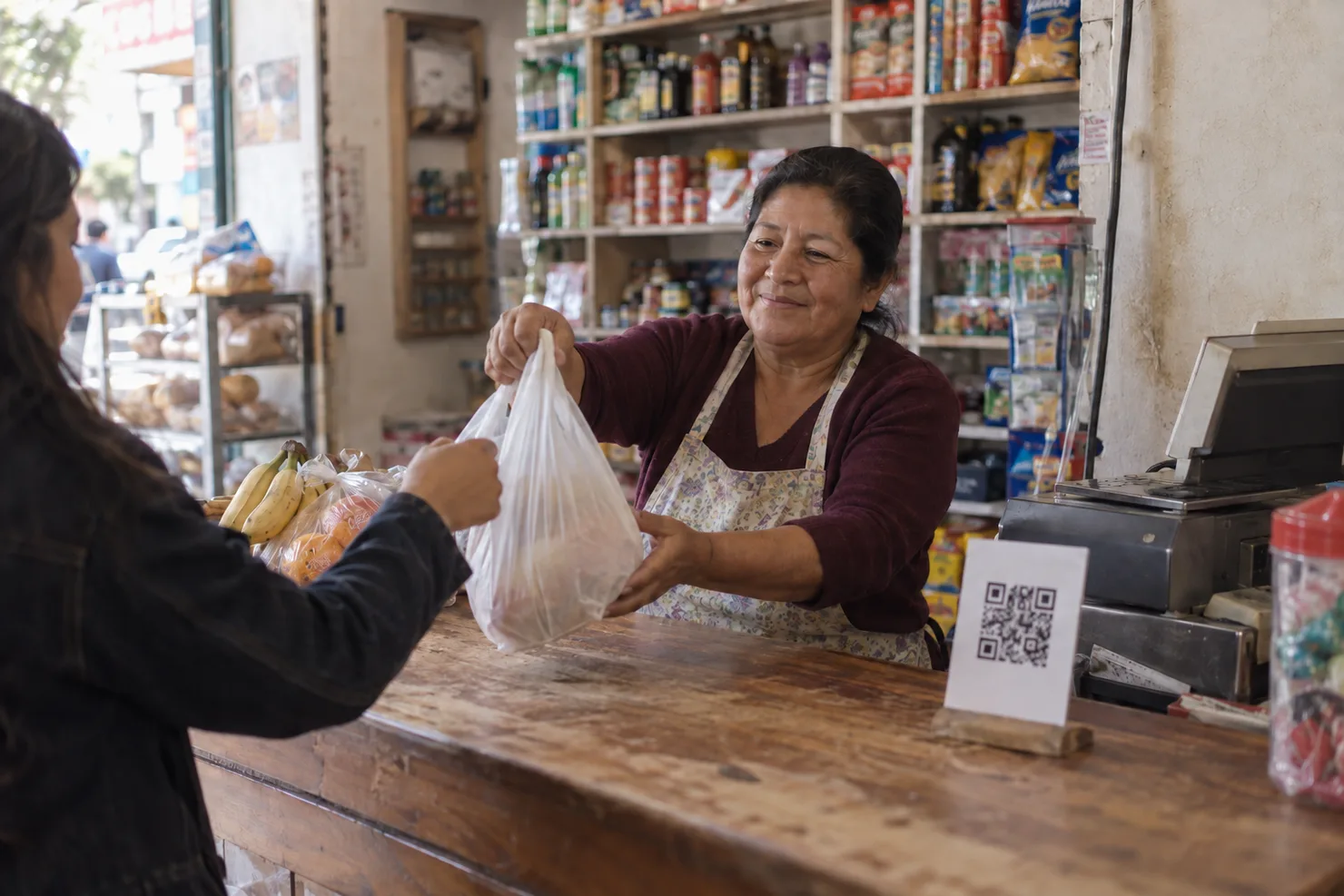 A bodega counter in Lima, a printed QR on a small stand beside the register, the shopkeeper handing a bag across