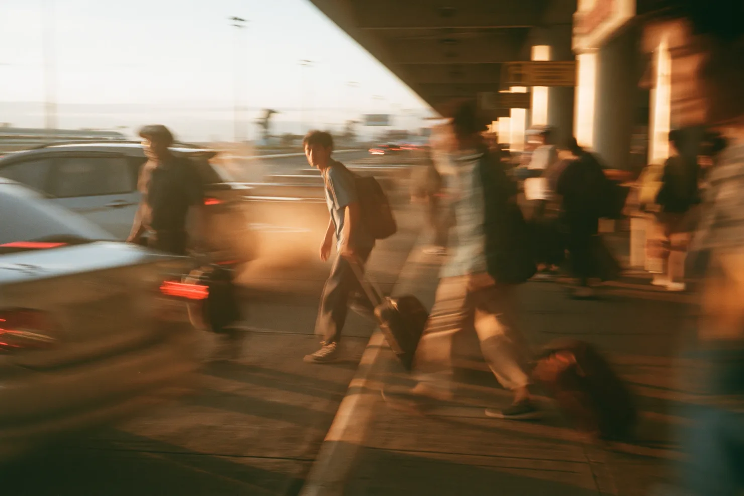 Travelers with rolling suitcases and backpacks stream along an airport arrivals curb in blurred warm motion at dusk