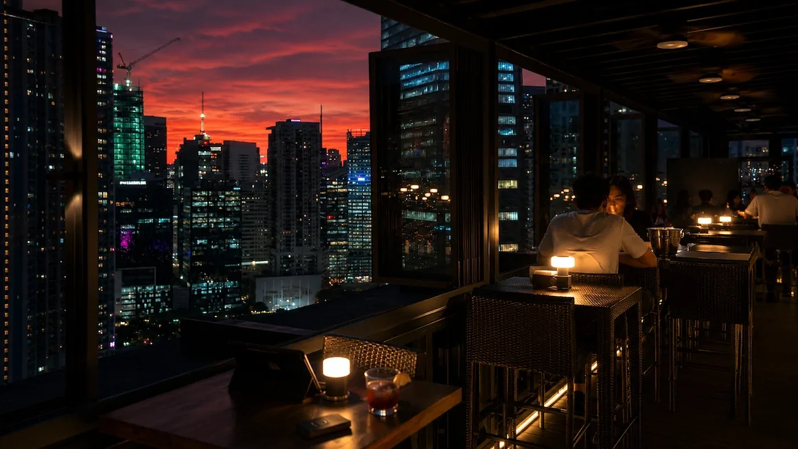 A rooftop bar at dusk — guests at high tables with small candle lamps looking out over a lit Manila skyline of towers under a fiery red sunset sky.