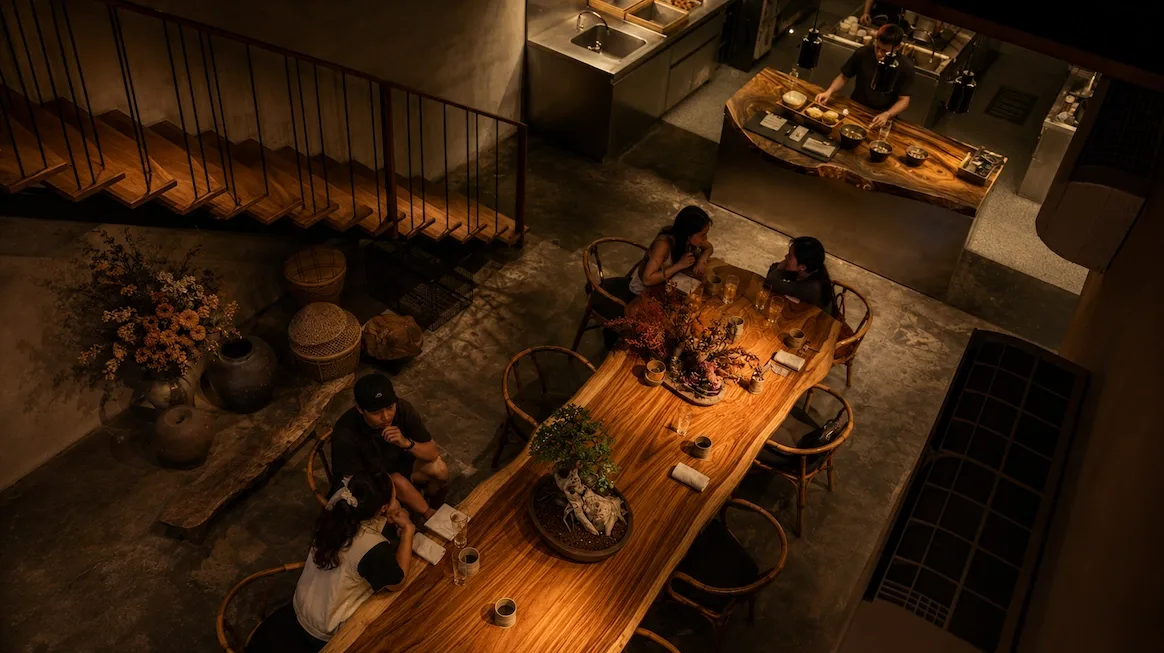 A high-angle view of Toyo Eatery's warm concrete-and-wood dining room — a long live-edge communal table with wishbone chairs and dried-flower arrangements, guests seated, and a chef plating at the open counter beyond.