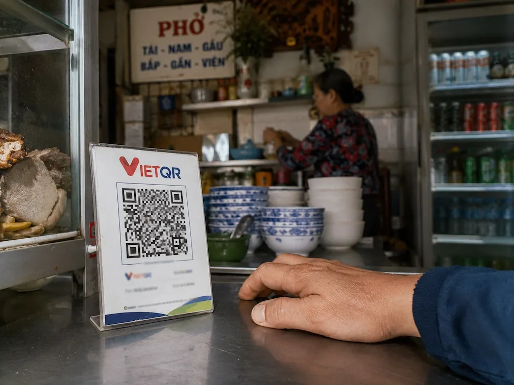 A VietQR payment standee on a Ho Chi Minh City pho-stall counter, a customer hand resting beside it