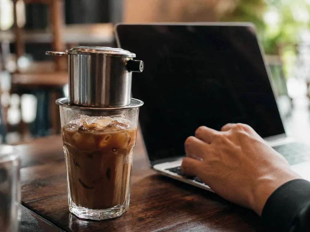 A glass of Vietnamese iced coffee with a phin filter beside an open laptop, a hand on the keys