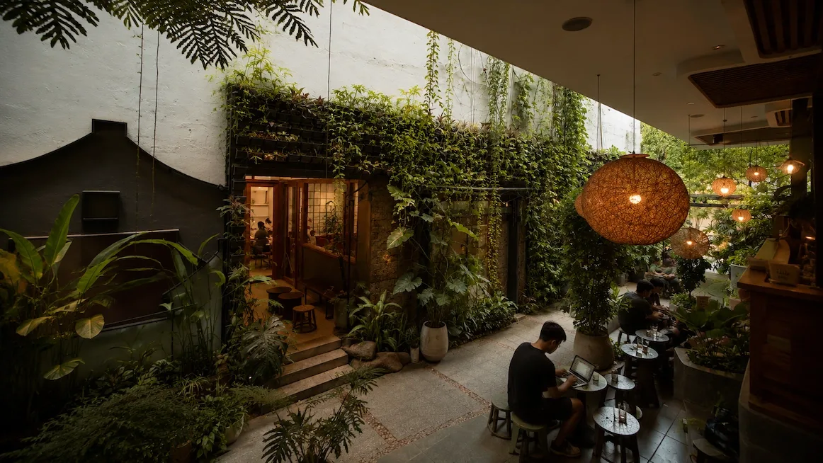 A plant-filled courtyard café — a person working on a laptop at a low table on a stool, ferns and vines climbing the white walls, and a woven rattan pendant light overhead.