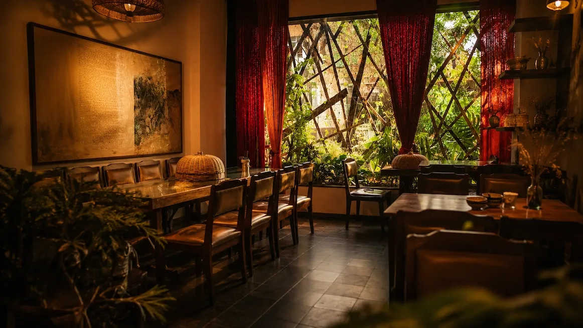 A warm, low-lit dining room at Cuc Gach Quan — wooden tables and leather chairs, woven rattan food covers, a large framed ink painting on the wall, and red curtains framing a window onto a green garden.