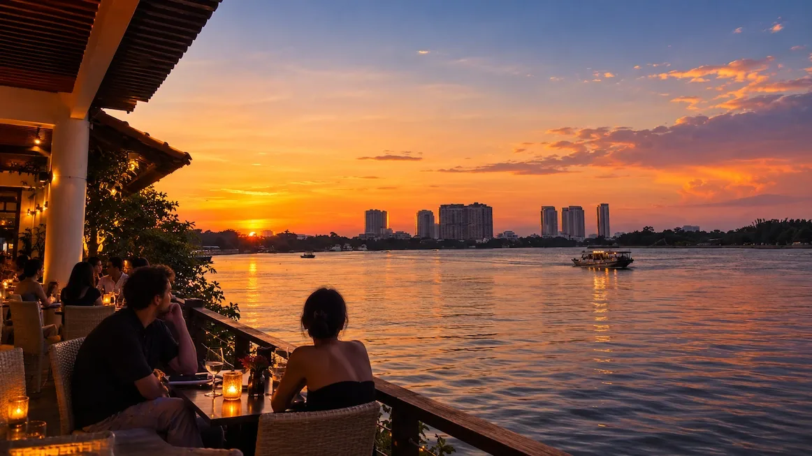 Diners at candlelit tables on a riverside terrace at sunset, looking out over the Saigon River as a boat passes and high-rises catch the last orange light across the water.