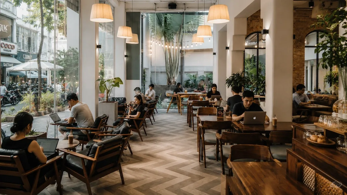Laptop users seated at wooden tables inside a bright, plant-filled Saigon cafe with pendant lamps and a small courtyard palm.