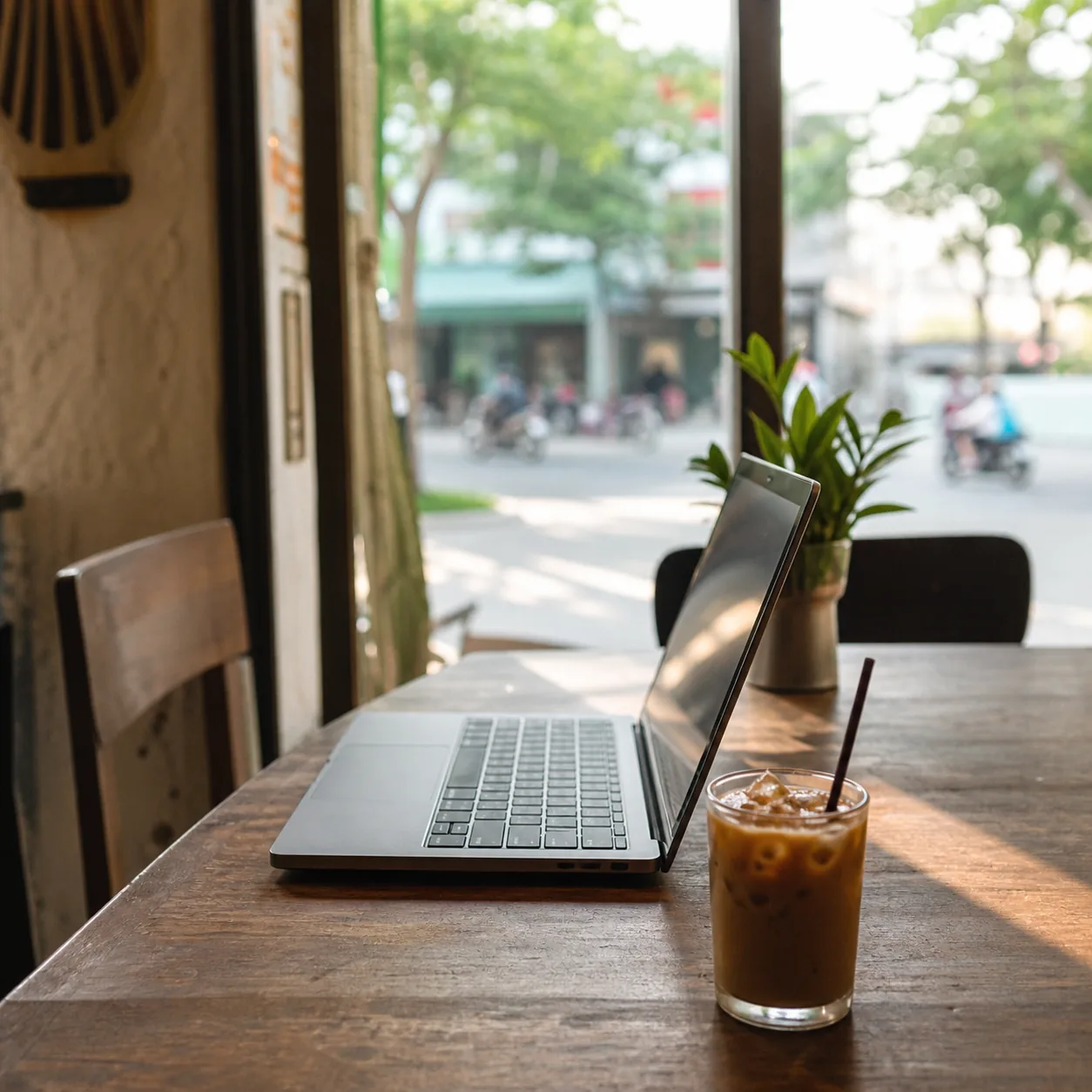 A laptop open on a cafe table in Da Nang with a glass of iced Vietnamese coffee beside it, morning light through the window