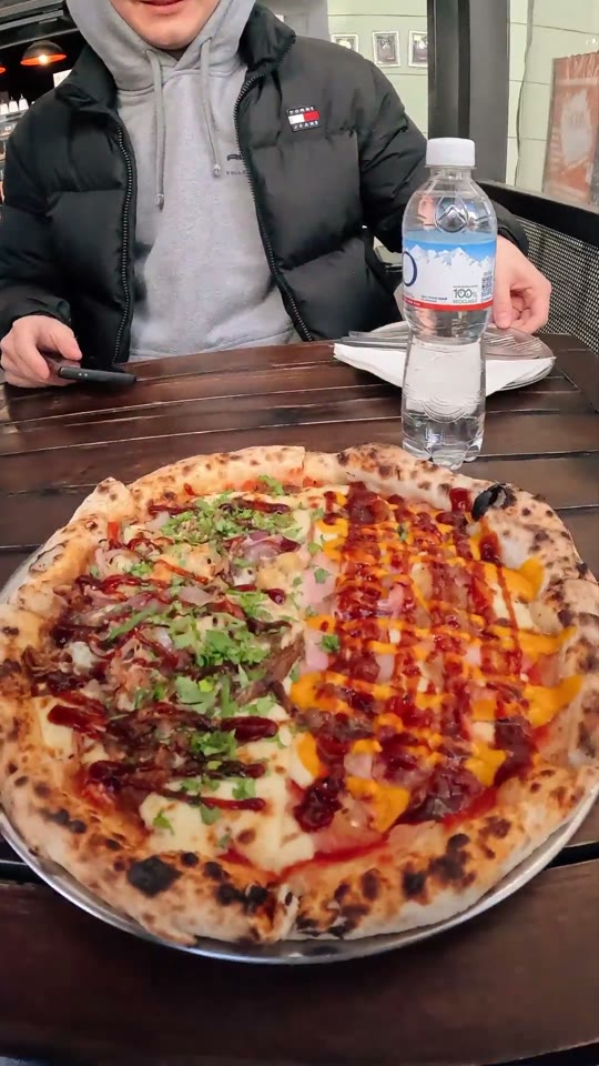 A blistered two-topping pizza on a metal tray at an outdoor Buenos Aires table, a diner in a puffer jacket and a water bottle behind it.