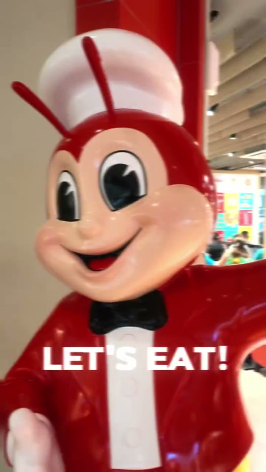 The red Jollibee mascot statue mid-wave with a 'let's eat' caption, inside a bright Manila mall food court.