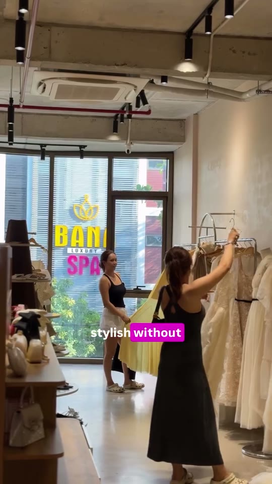 Two women browse a rack of pale dresses inside a Saigon clothing-rental boutique, one holding up a yellow gown by a bright window.